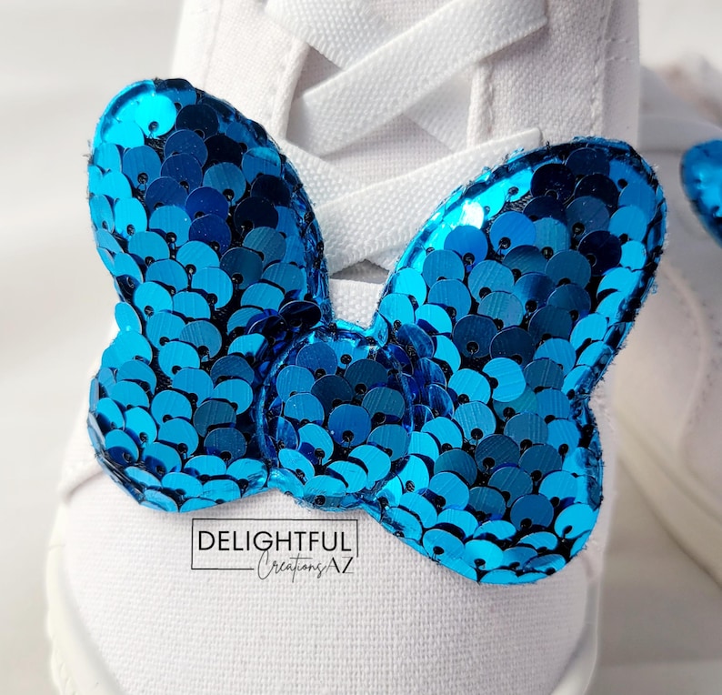 A close-up of a white sneaker with laces, showcasing a Minnie Mouse bow-shaped decoration attached to the front. The bow is adorned with shiny turquoise sequins, creating a vibrant and sparkling appearance. At the bottom of the image, the text "Delightful Creations AZ" emphasizes the branding, adding a personal and professional flair to the accessory.