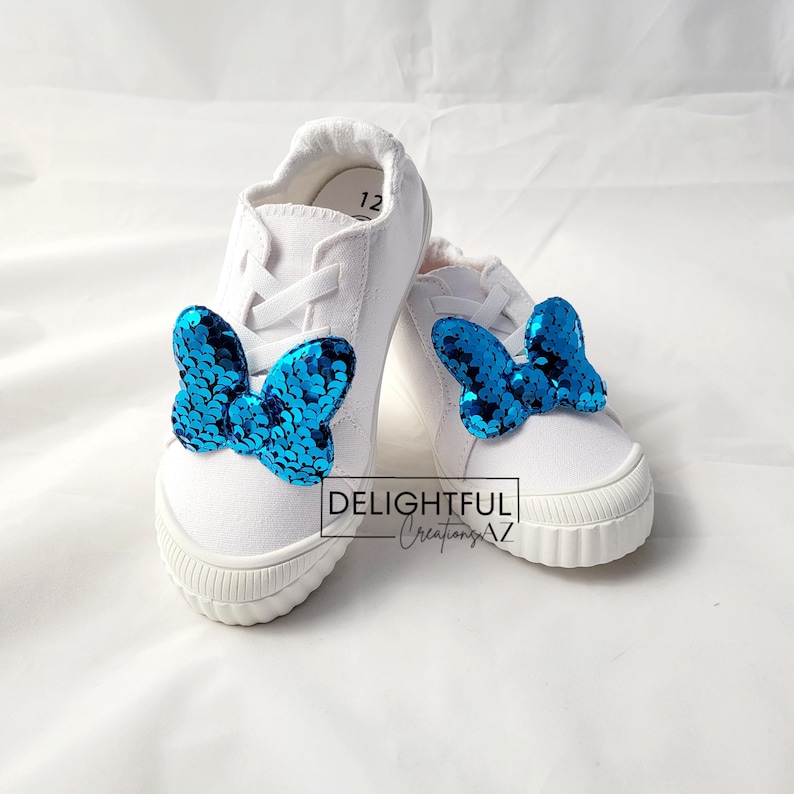 A pair of white sneakers featuring turquoise sequin Minnie Mouse bow-shaped decorations attached to the front. The shoes are displayed against a clean, white background, with the text "Delightful Creations AZ" prominently positioned in the center, emphasizing the brand identity. The design adds a playful and whimsical touch to the footwear.