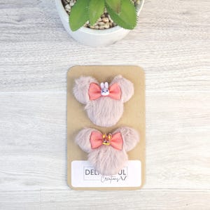 Puede incluir: Dos pinzas para el pelo rosas y esponjosas con forma de cabeza de rat&oacute;n, cada una con un lazo coral y un peque&ntilde;o adorno de personaje. Las pinzas se muestran en una tarjeta beige con el texto "DEL... Creations AZ". Una peque&ntilde;a planta est&aacute; en el fondo.