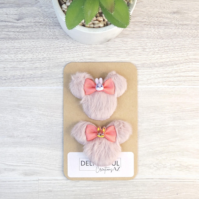 Puede incluir: Dos pinzas para el pelo rosas y esponjosas con forma de cabeza de rat&oacute;n, cada una con un lazo coral y un peque&ntilde;o adorno de personaje. Las pinzas se muestran en una tarjeta beige con el texto "DEL... Creations AZ". Una peque&ntilde;a planta est&aacute; en el fondo.