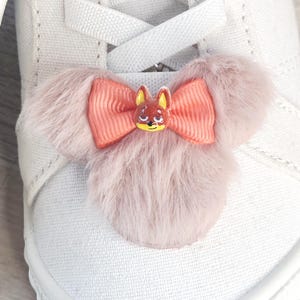 Puede incluir: Primer plano de un zapato de lona blanco con un adorno decorativo. El adorno presenta un dise&ntilde;o de piel rosa claro y esponjoso con un lazo color coral y una peque&ntilde;a cara de zorro pintada en el centro.