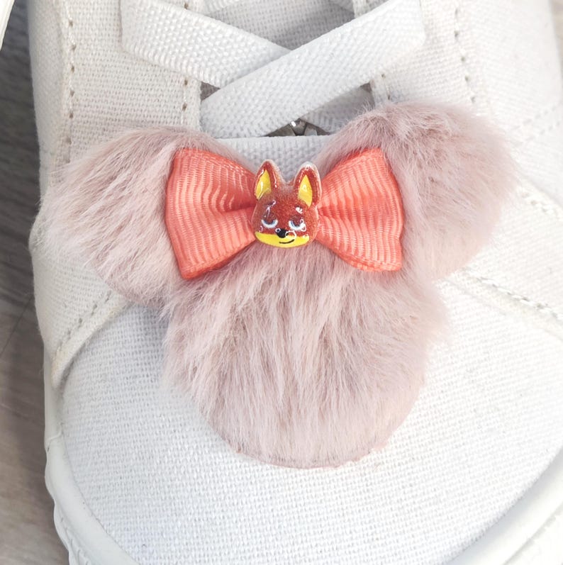 Puede incluir: Primer plano de un zapato de lona blanco con un adorno decorativo. El adorno presenta un dise&ntilde;o de piel rosa claro y esponjoso con un lazo color coral y una peque&ntilde;a cara de zorro pintada en el centro.