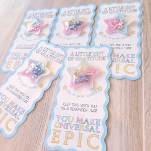 May include: Several star-shaped gifts with a clear, glitter-filled center are displayed on cards. The cards have a light blue border and text that reads "A Little Gift of Gratitude" and "You Make Universal Epic."