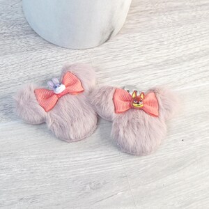 Puede incluir: Dos pinzas para el pelo con forma de orejas de rat&oacute;n rosa claro, con lazos coral y adornos de personajes. Una pinza tiene un conejito, la otra un zorro. Sobre una superficie de imitaci&oacute;n madera clara.