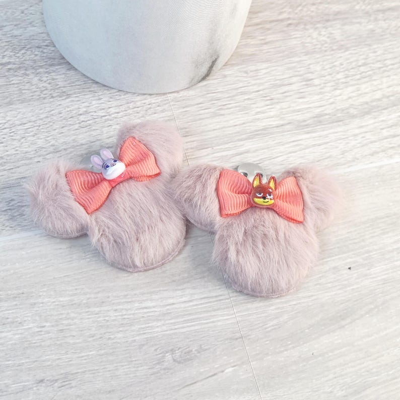 Puede incluir: Dos pinzas para el pelo con forma de orejas de rat&oacute;n rosa claro, con lazos coral y adornos de personajes. Una pinza tiene un conejito, la otra un zorro. Sobre una superficie de imitaci&oacute;n madera clara.
