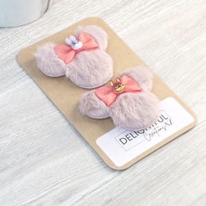 Puede incluir: Dos pinzas para el pelo rosas claras y esponjosas con forma de orejas de rat&oacute;n, cada una con un lazo coral y un peque&ntilde;o adorno de personaje. Las pinzas est&aacute;n sobre una tarjeta marr&oacute;n con el texto "DELIGHTFUL Creations AZ".