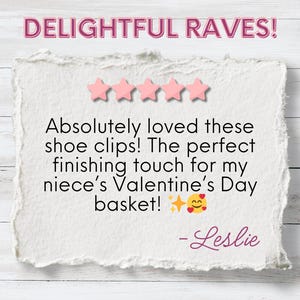 The image features a positive review titled "DELIGHTFUL RAVES!" displayed in bold pink text, accompanied by a five-star rating. The review reads: "Absolutely loved these shoe clips! The perfect finishing touch for my niece’s Valentine’s Day basket! ✨😍" and is signed by Leslie. The background has a clean white texture, giving the presentation a polished and cheerful vibe.