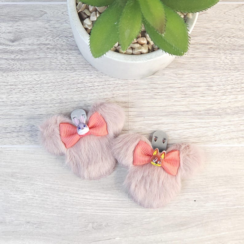 Puede incluir: Dos pinzas para el pelo rosas claras y esponjosas con forma de orejas de rat&oacute;n, cada una con un lazo coral y un dije de personaje. Una tiene un conejo, la otra un zorro. Sobre una superficie de vetas de madera clara.