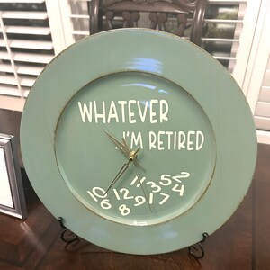 May include: A light blue round clock with gold numbers and hands. The clock is made from a repurposed plate and has the text "Whatever I'm Retired" written on it.