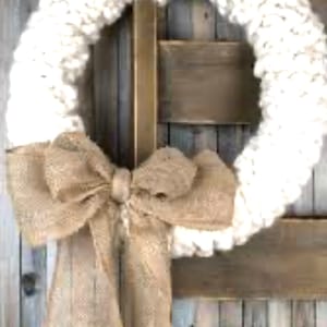 May include: A white knitted wreath with a burlap bow. The wreath is hanging on a wooden door.