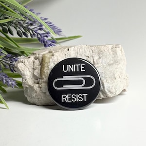 May include: A black button with the words "UNITE" and "RESIST" in white, along with a white paperclip graphic. The button is set against a light-colored rock and greenery.