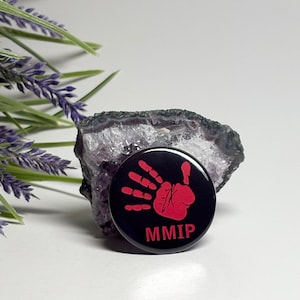 May include: A round, black button with a red handprint design and the letters "MMIP" in red. The button is set against a backdrop of a purple and gray geode and lavender sprigs.