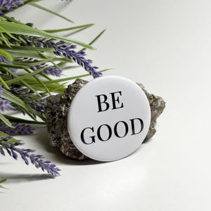 May include: A white button with the words "BE GOOD" in black, resting on a textured, gray stone. The button is round and centered. Green and purple foliage is in the background.