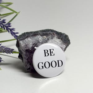 May include: A white button with the words "BE GOOD" in black lettering. The button is placed in front of a dark gray and purple geode. The background includes a sprig of purple flowers.