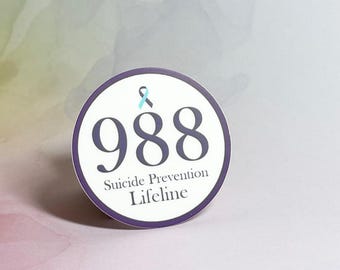 May include: A round sticker with a purple border and a white background. The sticker has a blue ribbon and the number 988 in black text. The text "Suicide Prevention Lifeline" is written in black text below the number.