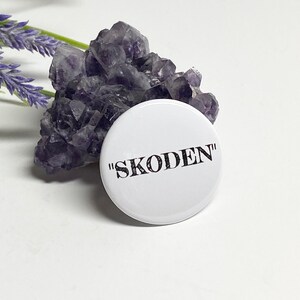 May include: A white button with the word "SKODEN" in black, set against a backdrop of a purple crystal cluster and a sprig of lavender. The button is round and the text is in a bold, sans-serif font.