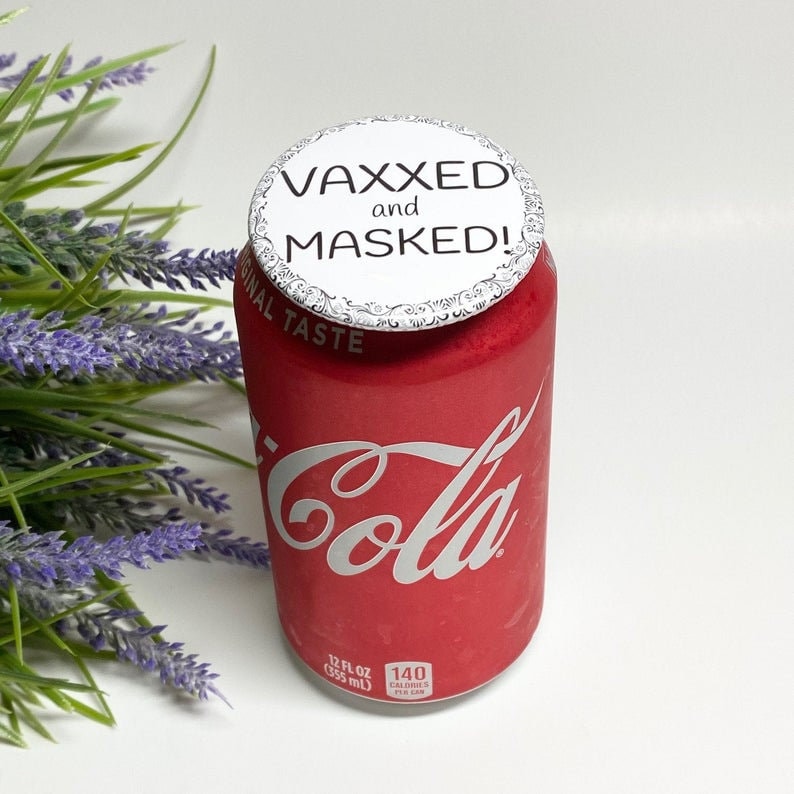 以下が含まれることがあります： 「VAXXED and MASKED!」と書かれた白い円形の蓋が付いた赤いコカ・コーラの缶。缶には銀色のコカ・コーラのロゴが入っています。缶は355mlです。