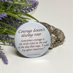 May include: A white button with a quote about courage. The quote reads, "Courage doesn't always roar. Sometimes courage is the little voice at the end of the day that says, 'I will try again tomorrow.'" - Mary Anne Radmacher.