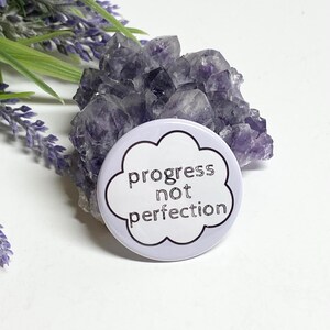 May include: White button with a purple background and a black outline of a cloud with the text "progress not perfection" inside.