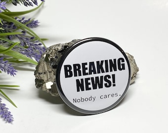 May include: A round, black and white button with the text "BREAKING NEWS!" in bold, black letters. Below the text is the phrase "Nobody cares." The button is resting on a piece of pyrite and is next to a sprig of lavender.