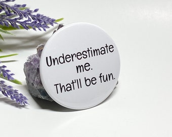 May include: White round button with black text that reads "Underestimate me. That'll be fun."
