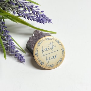 May include: A round, cream-colored button with a blue floral border. The button has the words "faith over fear" written in blue cursive.