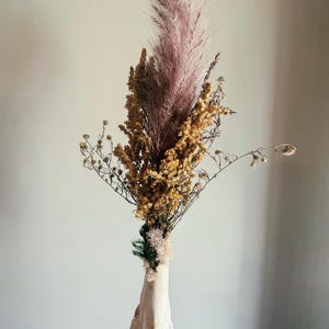 May include: A dried floral arrangement featuring a large, feathery, pink pampas grass plume, surrounded by smaller dried yellow and brown flowers. The arrangement is held in a cream-colored, bone-shaped vase.