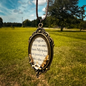 May include: An oval pendant on a brown cord featuring an ornate bronze-toned metal frame. The center contains printed text with the visible words my legs, my ass and face, my puss, on panties, and a small white bone fragment with dried plant elements.