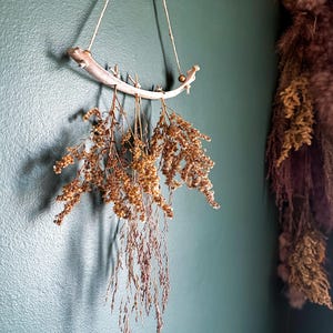 May include: A wall hanging decoration featuring a curved, light-colored wooden branch suspended by a thin twine cord. Several bundles of dried, golden-brown wildflowers and grasses are tied to the branch, hanging downward in a cluster.