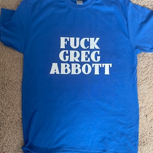 Puede incluir: Una camiseta azul real con el texto "FUCK GREG ABBOTT" impreso en blanco.