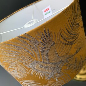 Puede incluir: Una pantalla de l&aacute;mpara dorada con un dise&ntilde;o de gr&uacute;a negro y plateado. La pantalla de la l&aacute;mpara est&aacute; hecha de tela y tiene un forro blanco. La etiqueta en la pantalla de la l&aacute;mpara dice "Hand Made Lampshades".