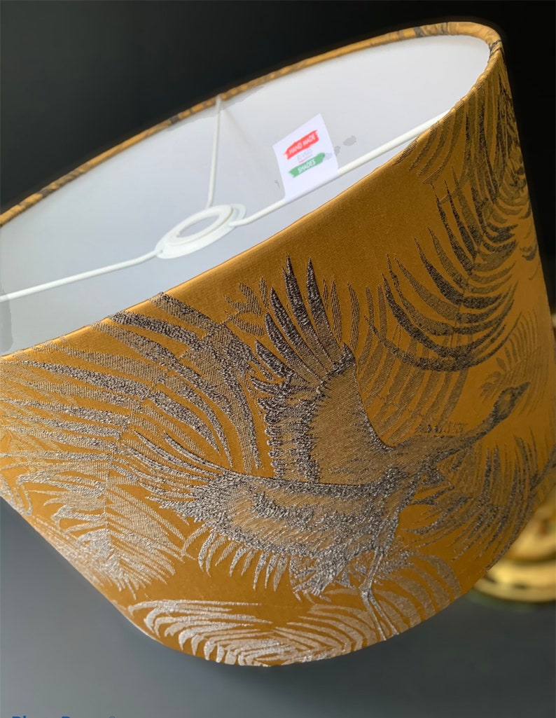 Puede incluir: Una pantalla de l&aacute;mpara dorada con un dise&ntilde;o de gr&uacute;a negro y plateado. La pantalla de la l&aacute;mpara est&aacute; hecha de tela y tiene un forro blanco. La etiqueta en la pantalla de la l&aacute;mpara dice "Hand Made Lampshades".