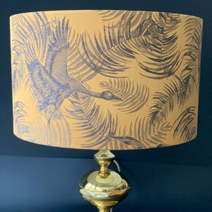 Puede incluir: Una l&aacute;mpara de mesa dorada con una pantalla amarilla con un patr&oacute;n de hojas tropicales en blanco y negro. La pantalla tiene una silueta de p&aacute;jaro volando a trav&eacute;s de las hojas.