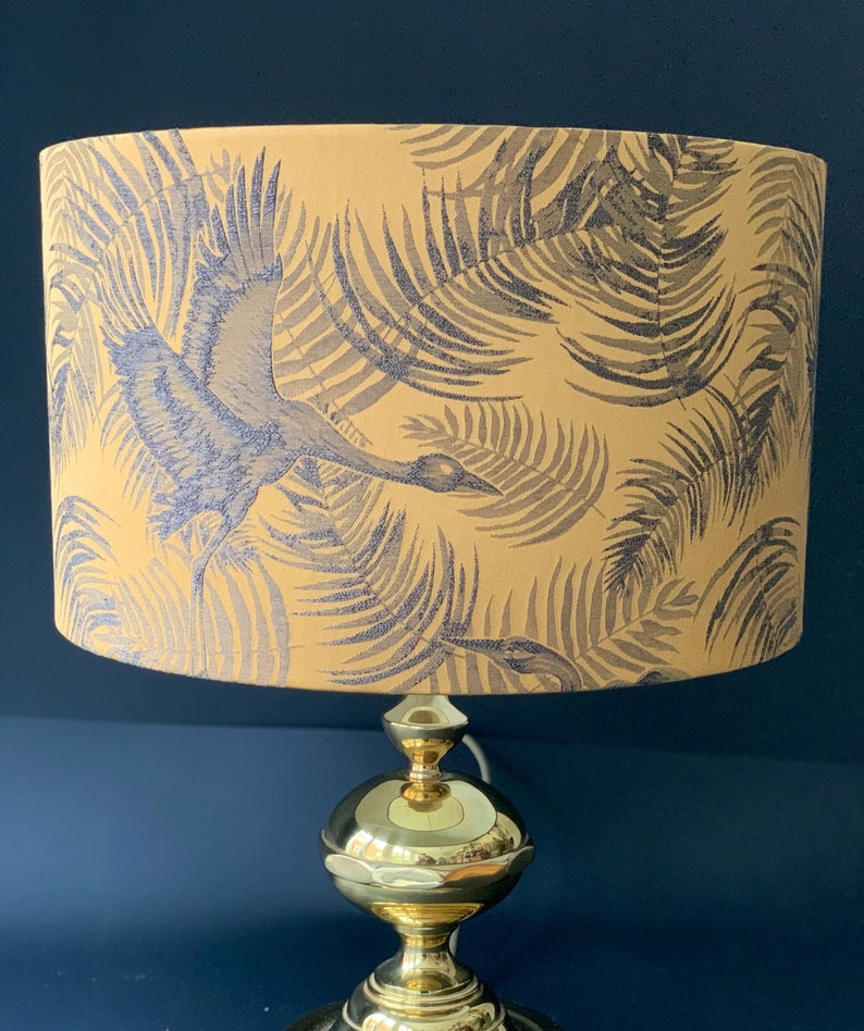Puede incluir: Una l&aacute;mpara de mesa dorada con una pantalla amarilla con un patr&oacute;n de hojas tropicales en blanco y negro. La pantalla tiene una silueta de p&aacute;jaro volando a trav&eacute;s de las hojas.