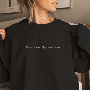 以下が含まれることがあります： 白い文字で「Please be nice. My tummy hurts.」とプリントされた黒いスウェットシャツ