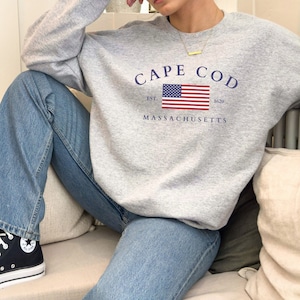 Puede incluir: Una sudadera gris claro con las palabras "CAPE COD" en azul marino arqueadas sobre un gráfico de la bandera estadounidense. Debajo de la bandera están las palabras "EST. 1620" y "MASSACHUSETTS" en azul marino. La sudadera se combina con vaqueros azules.
