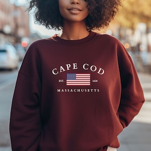 May include: Burgundy sweatshirt with a white graphic print that says "CAPE COD EST. 1620 MASSACHUSETTS" and features a small American flag.