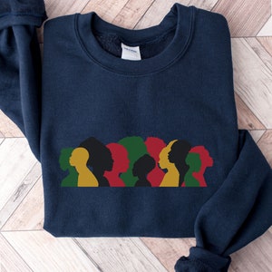 Op de afbeelding: Een donkerblauw sweatshirt met een afbeelding van een rij zwarte silhouetten van mensen met verschillende kapsels. De silhouetten zijn omrand met geel, rood en groen.