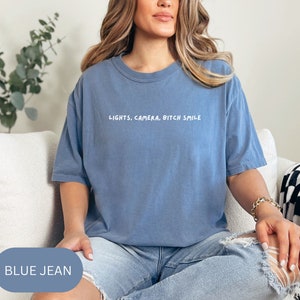 Könnte beinhalten: Ein Oversize-T-Shirt in Jeansblau mit dem Aufdruck "LIGHTS, CAMERA, BITCH SMILE" in Weiß.