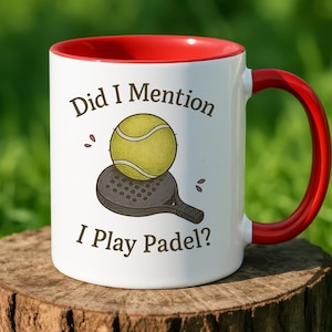 May include: White ceramic mug with a red interior and handle. The mug features the text "Did I Mention I Play Padel?" along with an illustration of a padel ball and racket. The mug is sitting on a wooden stump.