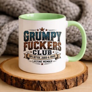 Puede incluir: Una taza de cerámica blanca con interior y asa de color verde claro. El frente presenta el texto GRUMPY FUCKERS CLUB en letras desgastadas azules y marrones, con el subtítulo 100% BITTER, JADED & PROUD y LIFETIME MEMBER, decorado con estrellas.
