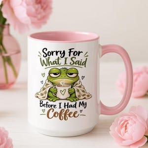 Puede incluir: Una taza de cerámica blanca con interior y asa de color rosa, que presenta un gráfico de una rana verde envuelta en una manta sosteniendo una taza de café. El texto dice: Sorry For What I Said Before I Had My Coffee, con pequeños corazones verdes.