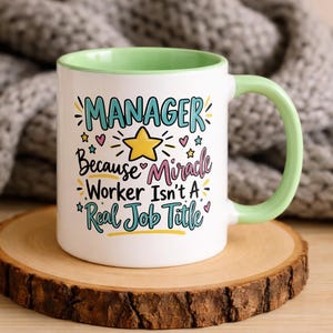 Könnte beinhalten: Ein weißer Keramikbecher mit hellgrünem Innenraum und Henkel. Auf der Vorderseite steht der Text "MANAGER Because Miracle Worker Isn't A Real Job Title" in Blau, Schwarz und Pink, verziert mit einem gelben Stern sowie kleinen bunten Herzen und Sternen.