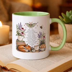 May include: A white ceramic mug with a light green interior and handle, featuring a printed illustration of a sleeping fox, a moth, flowers, and a stack of books. The text reads: May your days be filled with slow mornings, warm tea, forest walks, fresh bread, blooming flowers, crackling fires, starry skies, and people who make your soul feel at home.