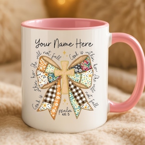 Può includere: Tazza in ceramica bianca con interno e manico rosa. La tazza presenta un design a fiocco con una croce al centro e il testo "Your Name Here" e "God is within her, she will not fall" in scrittura nera. Il design include motivi floreali e a quadri.
