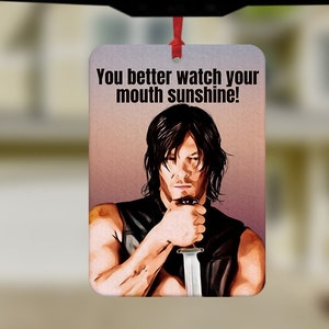 May include: A car air freshener featuring a man holding a knife. The text on the air freshener reads "You better watch your mouth sunshine!"