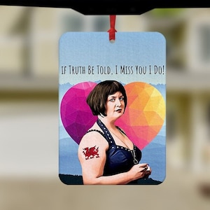 Pode incluir: Um aromatizador de carro azul com o rosto de uma mulher e uma tatuagem de dragão vermelho. O texto no aromatizador diz "If truth be told, I miss you I do!"