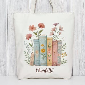 Può includere: Borsa tote color crema con un'illustrazione ad acquerello di libri e fiori. I libri sono in vari colori pastello, con dettagli floreali. Il nome "Charlotte" è scritto in basso. La borsa ha manici color crema.
