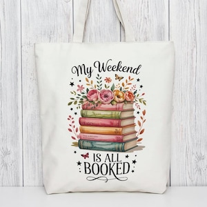 Pode incluir: Saco tote branco sujo com um design com uma pilha de livros adornados com flores e o texto "My Weekend Is All Booked". Os livros têm lombadas coloridas, e o arranjo floral inclui flores rosa, laranja e amarelas.