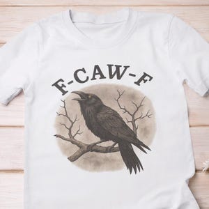 Op de afbeelding: Wit T-shirt met een afbeelding van een zwarte kraai op een tak, omringd door kale takken, met de tekst "F-CAW-F" erboven. De afbeelding bevat ook witte sneakers en een geruit overhemd. De achtergrond is lichtgekleurd hout.